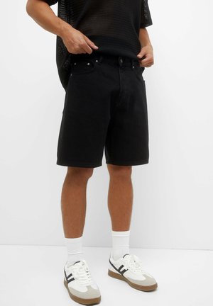 Person iført sorte shorts, hvide sokker, hvide sneakers med sorte striber og en sort net-top, justerer deres shorts taljebånd.