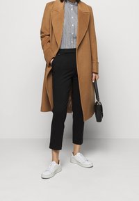 Brun ullrock med skräddarsydd design, svarta byxor och vita sneakers. Utrustningen inkluderar en randig skjorta under och en svart handväska.