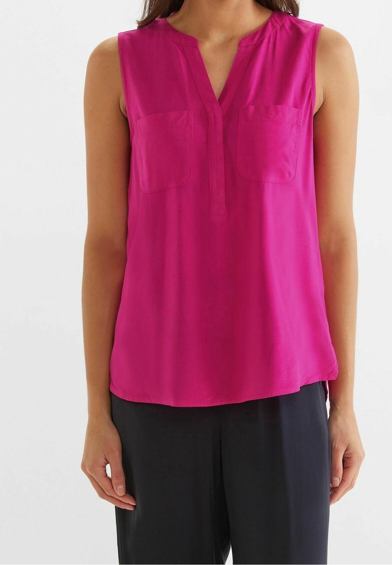 Blouse rose vif sans manches avec deux poches poitrine et un col en V fendu, portée avec un pantalon noir ample.