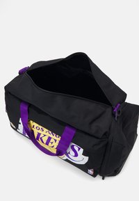 Fekete sporttáska tartós anyagból, lila fogantyúkkal és cipzárral. Elöl a "Los Angeles Lakers" logója sárga és fehér színben látható.