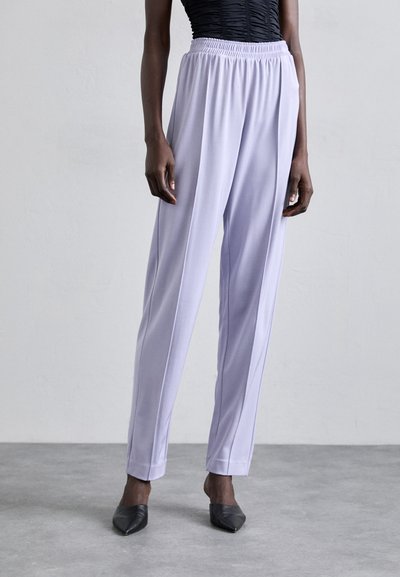 Pantalons lavande clairs en tissu lisse, dotés d'une taille élastique et d'une coupe droite avec des plis. Associés à des chaussures noires à bouts pointus.