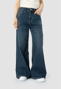 Cilvēks ģērbjas tumši zilos brīvajos biksēs un baltās kedās, ar vienu roku kabatā un daļēji redzamu baltu augšdaļu.