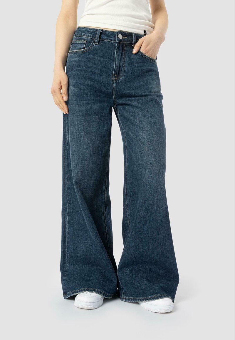 Cilvēks ģērbjas tumši zilos brīvajos biksēs un baltās kedās, ar vienu roku kabatā un daļēji redzamu baltu augšdaļu.