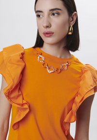 Camiseta naranja con mangas rizadas, que presenta un collar de cadena enlazada en tonos claros y naranjas, sobre un fondo blanco liso.