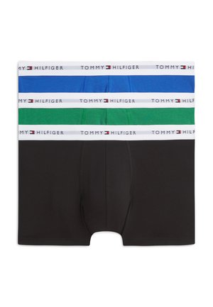 Ung mand står barfodet, uden trøje, iført blå boxershorts med en hvid linning med et brandlogo, mod en ens hvid baggrund.