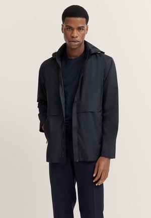 Schwarze wasserdichte Jacke mit Kapuze, ausgestattet mit einem Frontreißverschluss, zwei Fronttaschen und einer lockeren Passform. Glatte Textur und minimalistisches Design.
