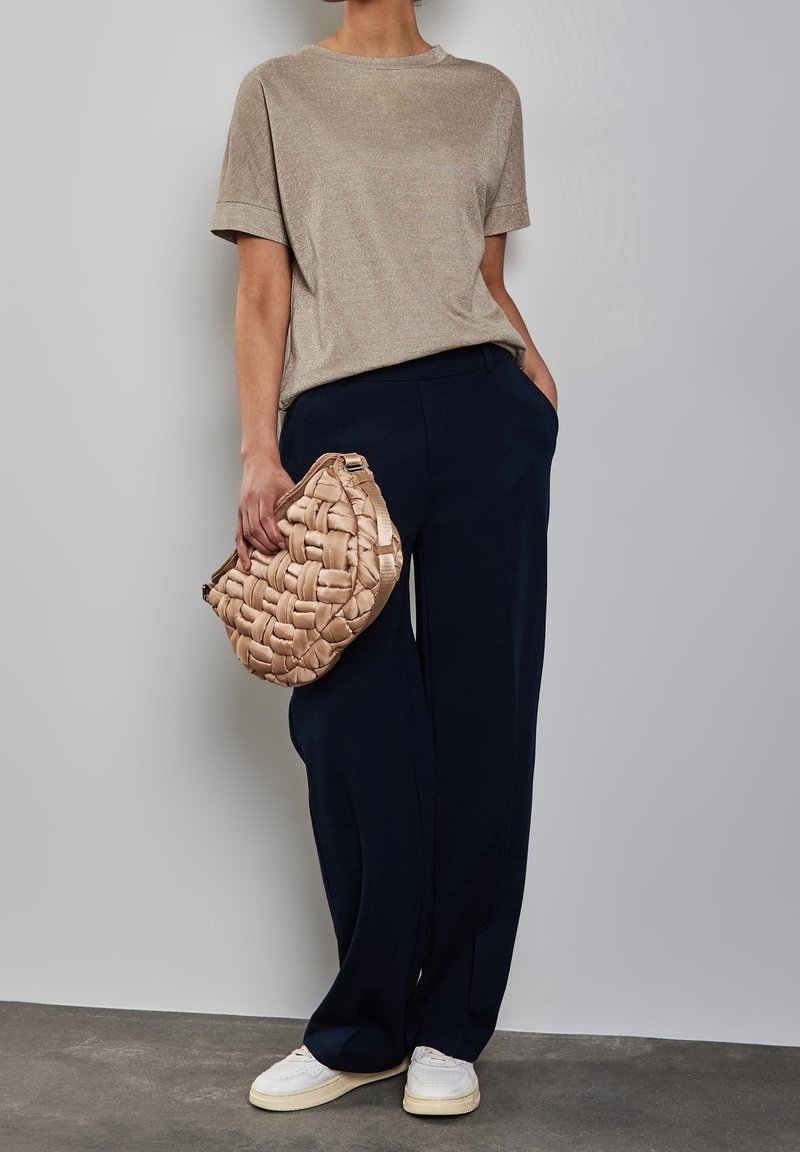 Person som bär en skimrande beige topp, marinblå vida byxor, vita sneakers, håller en vävd beige clutchväska, står mot en grå vägg.