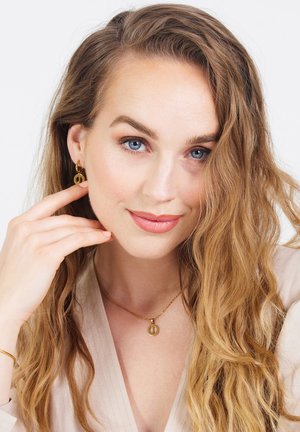 Goldene, runde Ohrringe und eine passende Anhängerkette. Welliges, schulterlanges Haar und neutrale Kleidung. Glatte Haut und dezentes Make-up.