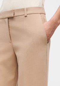 Μπεζ παντελόνι καλοραμμένο με πλαϊνή τσέπη, θηλιά για ζώνη και ένα χέρι μερικώς μέσα στην τσέπη, πάνω σε μονόχρωμο φόντο.