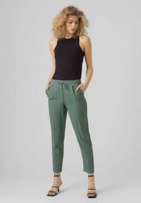 Zwarte mouwloze top gecombineerd met lichtgroene tapered broeken met een trekkoord in de taille en zijzakken. Model draagt zwarte sandalen met bandjes.