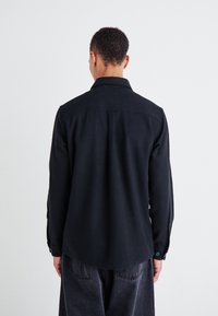 Chemise noire à manches longues au design minimaliste, avec un col et des poignets boutonnés. Tissu texturé et détail de l'empiècement du dos droit.