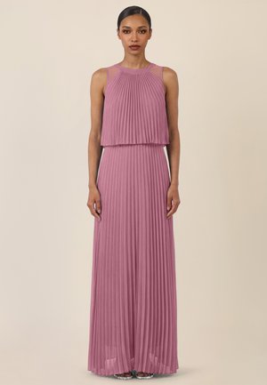 Frau steht und trägt ein ärmelloses maxilanges Kleid in Mauve mit plissiertem Rock und mehrlagigem Oberteil vor einem hellbeigen Hintergrund.