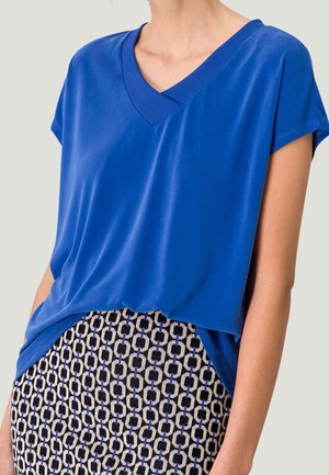 Kobieta w jasnoniebieskiej bluzce z krótkim rękawem i dekoltem w kształcie litery V, wpuszczonej w spódnicę z geometrycznym wzorem w kolorach czarnym, białym i niebieskim.