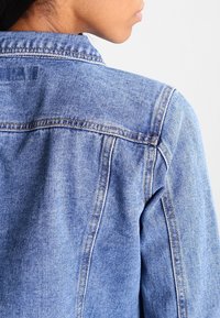 Jasnoniebieska kurtka dżinsowa o klasycznym kroju z widocznymi szwami, kołnierzem typu spread oraz lekko teksturowaną powierzchnią.