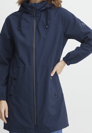 Femme portant une veste à capuche bleu marine avec fermeture éclair à l'avant, poignets élastiques et poches à boutons-pression, debout avec une main sur la hanche.