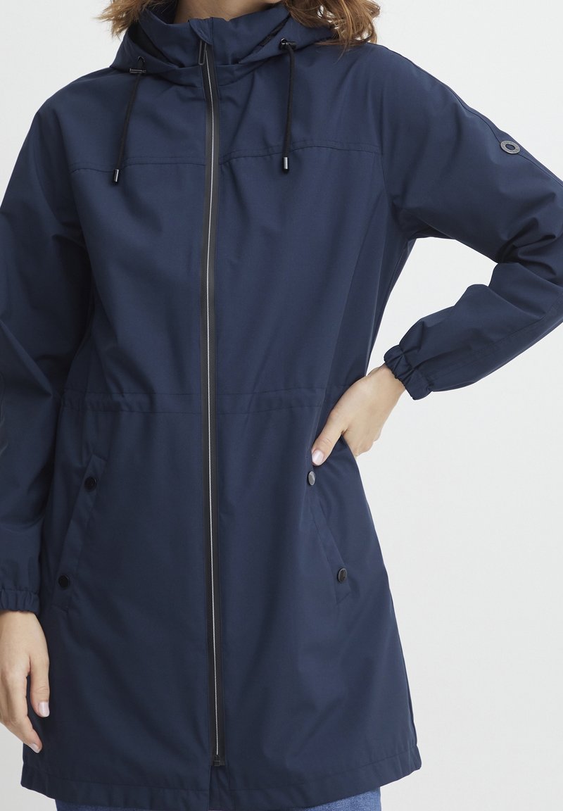 Femme portant une veste à capuche bleu marine avec fermeture éclair à l'avant, poignets élastiques et poches à boutons-pression, debout avec une main sur la hanche.