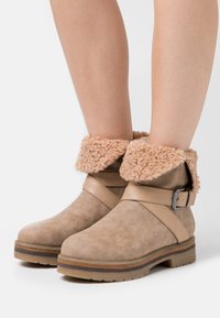 Ruske ante suede ankle boots s fluffy bež rubom, s akcentom od metalnog remena i ravnom gumiranom potplatom s teksturiranim detaljima.