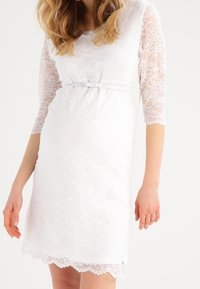 Robe en dentelle blanche avec des manches trois-quarts, un buste ajusté et une ceinture en satin à la taille. Ourlet festonné et détails en dentelle florale.