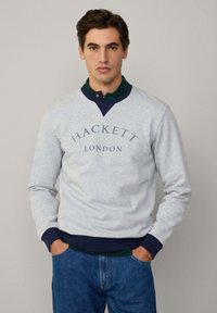 Sudadera gris con detalles en azul marino, con el logo de "HACKETT LONDON", puños acanalados y un ajuste casual, combinada con una camisa verde oscura debajo.