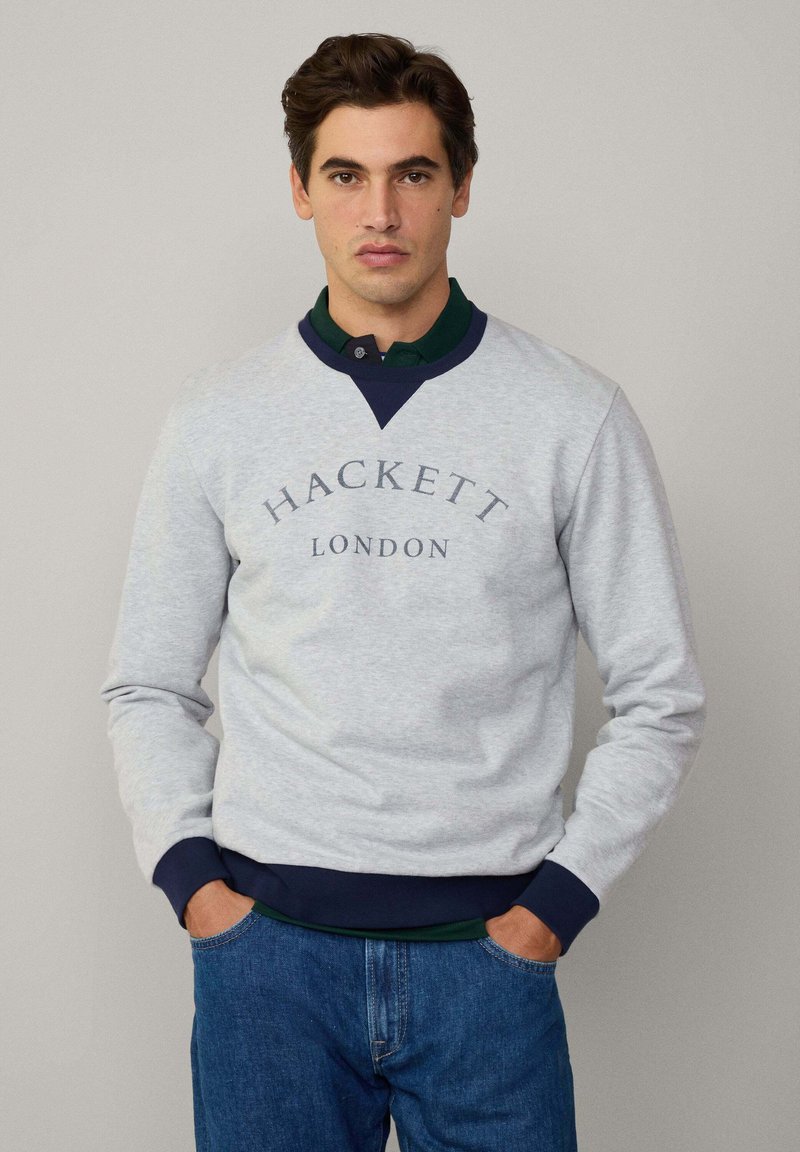 Sudadera gris con detalles en azul marino, con el logo de "HACKETT LONDON", puños acanalados y un ajuste casual, combinada con una camisa verde oscura debajo.
