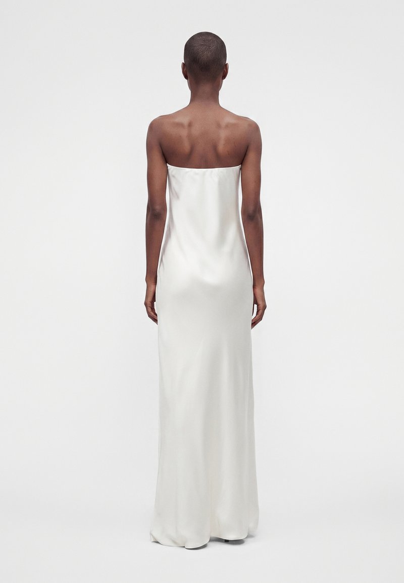 Personne aux cheveux courts portant une longue robe blanche en satin sans bretelles, debout, dos à la caméra, sur un fond uni.