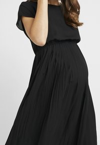 Mustapleated raskausmekko, jossa on lyhyet hihat, kerätty vyötärö, sileä pinta ja valuva kangas, joka laskeutuu vatsan päälle.