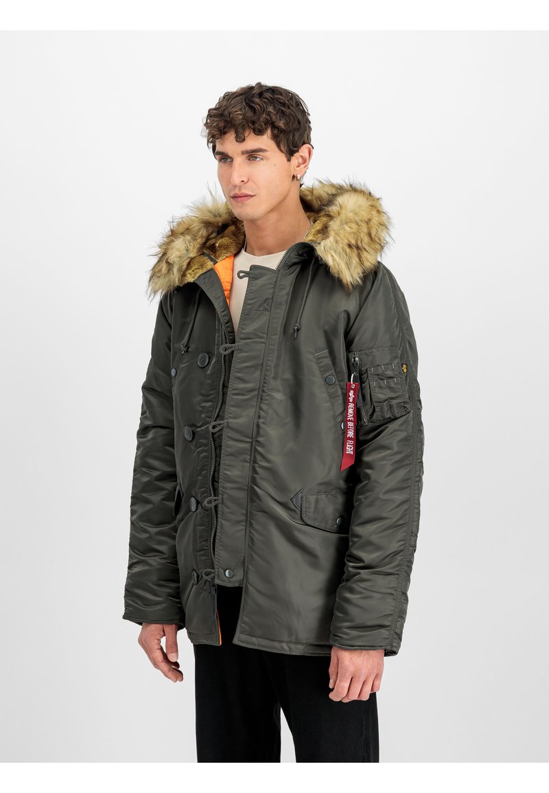Chaqueta aislante de color verde oliva con capucha de pelo sintético, cierre de cremallera y botón, bolsillos laterales y una etiqueta roja. Textura suave y diseño cálido.