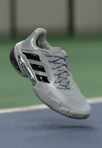 Grå sportsko med sorte striber og et indviklet således design, svævende over en blå og grøn sportsbane.
