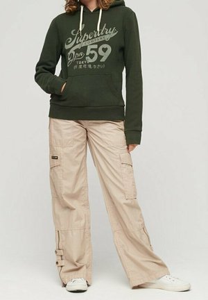 Personne portant un sweat à capuche Superdry vert foncé avec des cordons blancs, un pantalon cargo beige avec poches latérales et des baskets blanches, debout devant un fond uni.