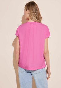 Blusa a maniche corte di colore rosa pastello con una texture liscia e scollatura rotonda, caratterizzata da un sottile dettaglio arricciato sul retro. Abbinata a jeans azzurri.