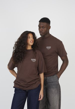 Due giovani adulti che indossano magliette marroni con la scritta "Legacy"; la donna ha capelli ricci e jeans scuri, l'uomo è dietro di lei con jeans grigi scoloriti.