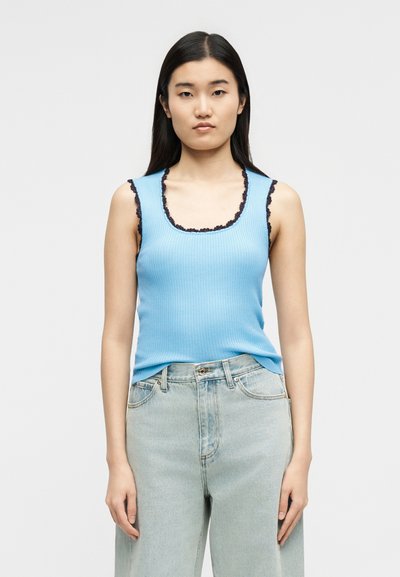 Νεαρή γυναίκα με μακριά σκούρα μαλλιά, φοράει αμάνικο γαλάζιο τοπ με ρίγες και μαύρη δαντέλα στις άκρες, καθώς και ανοιχτόχρωμα τζιν, στέκεται μπροστά σε λευκό φόντο.