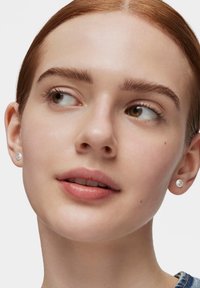 Mujer joven de piel pálida y cabello rojo mira de lado, llevando pendientes de botón con un pequeño emblema sobre un fondo blanco.