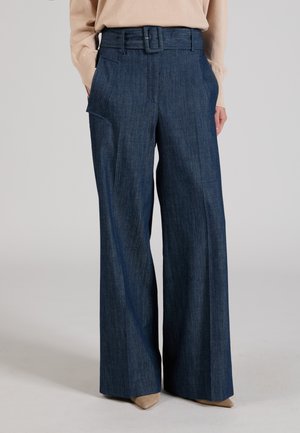 Hoch taillierte, weit geschnittene dunkelblaue Jeanshose mit passendem Gürtel und Seitentaschen, kombiniert mit beigem Langarmshirt und beigen spitzen Schuhen.