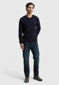 Marineblauer V-Ausschnitt-Pullover aus weichem Strick, kombiniert mit dunklen Jeans. Das Model steht mit den Händen in den Taschen und trägt graue Schuhe. Einfaches und modernes Design.