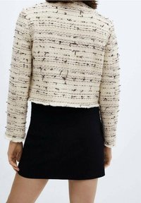 Vue de dos d'une personne portant une veste en tweed texturée crème avec des taches noires et une jupe courte noire sur un fond blanc.