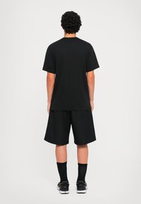 Άνδρας με σγουρά μαλλιά που στέκεται με την πλάτη στραμμένη, φορώντας μαύρο μπλουζάκι, μαύρες σορτς μέχρι το γόνατο, μαύρες κάλτσες και μαύρα αθλητικά παπούτσια.