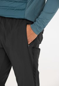 Un pantalon de performance noir avec une poche zippée, associé à un haut à manches longues teal en tissu lisse et léger.