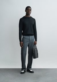 Muž v čiernom svetri, sivých nohaviciach a čierno-bielych mokasínach, držiaci čiernu aktovku, stojaci pred jednoduchým svetlým pozadím.