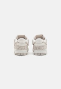 Beige és fehér Nike alacsony szárú sportcipők párja, szürke foltos mintázattal, a hátulról látható, fehér háttér előtt.