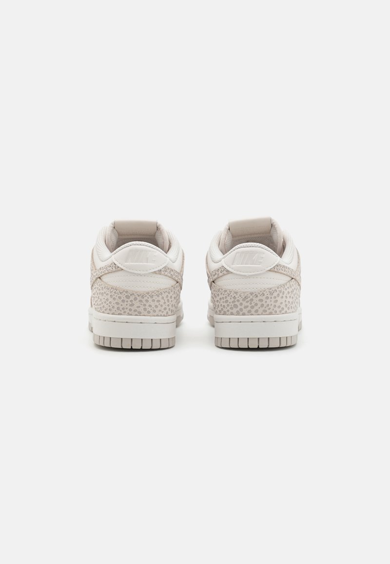 Par de zapatillas Nike de bajo corte en beige y blanco con un patrón moteado gris, vistas desde atrás sobre un fondo blanco.