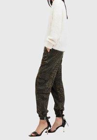 Weißer, flauschiger Pullover, gemusterte, dunkelgrüne Cargo-Hose mit Taschen und schwarze High-Heelsandaletten mit Knöchelriemen und goldenen Akzenten.