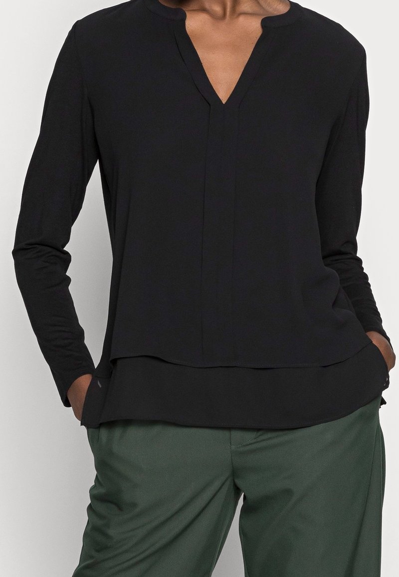 Maglia nera a maniche lunghe con scollo a V, morbida al tatto e con orlo a strati. Abbinata a pantaloni verdi, con tasche visibili su entrambi i lati.