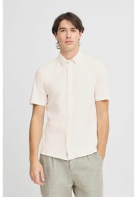 Kortermet skjorte med knappestolpe i lys beige stoff, med standard krage og en tettsittende design, kombinert med lyse grå bukser. Glatt tekstur.