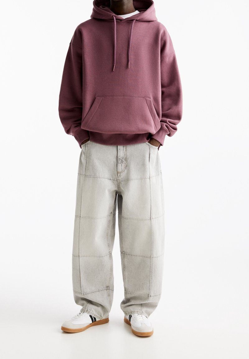 Personne portant un sweat à capuche bordeaux et un pantalon ample gris clair avec de grandes coutures apparentes, ainsi que des baskets blanches à semelles en gomme.
