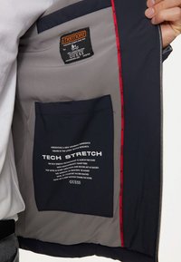 L'interno della giacca navy presenta un rivestimento grigio, una tasca nera con testo bianco e un'etichetta di marca. Cuciture di accento rosse lungo la cerniera.
