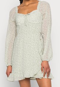 Vestido floral verde claro con mangas largas transparentes, escote cuadrado y un dobladillo con volantes. Presenta un lazo en la cintura y un sutil patrón texturizado.