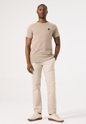 Beige gestricktes Kurzarmshirt mit Rundhalsausschnitt und kleinem Logo, kombiniert mit hellbeigen Hosen und beigen Sneakern mit braunen Akzenten.