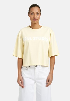 Woman with pulled-back hair wears pale yellow oversized T-shirt with "SOUL STUDIO" text and white high-waisted jeans against plain background.