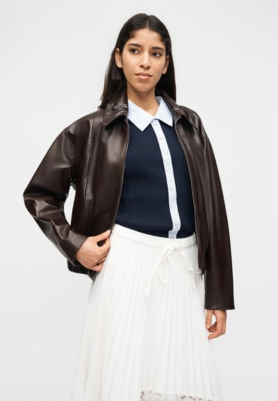 Γυναίκα που φοράει καφέ σκούρο δερμάτινο μπουφάν πάνω από μπλε ναυτικό πουκάμισο με λευκό γιακά, συνδυασμένο με άσπρη πλισέ φούστα, στέκεται μπροστά σε απλό φόντο.